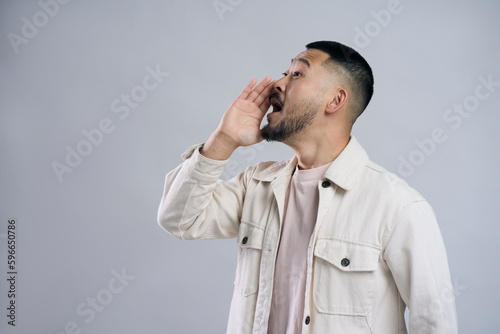 Asian man holding hand near face, screaming loud about sale, looking away isolated on gray background. Promotion, shopping concept