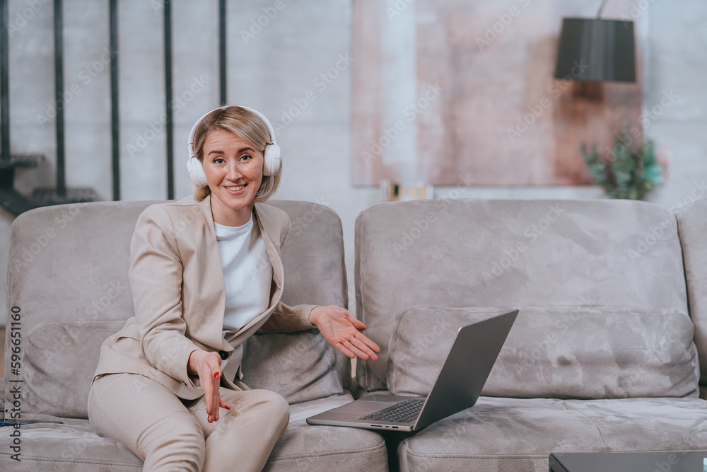 Blonde American young woman in beige suit and headphones sitting on cozy couch using laptop looks at camera spreads hands in surprise. Successful caucasian teacher at remote lesson.