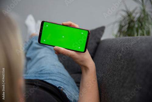 Back view of young europen woman who is using chroma key smartphone screen.She is sitting on the sofa in her cozy living room. photo
