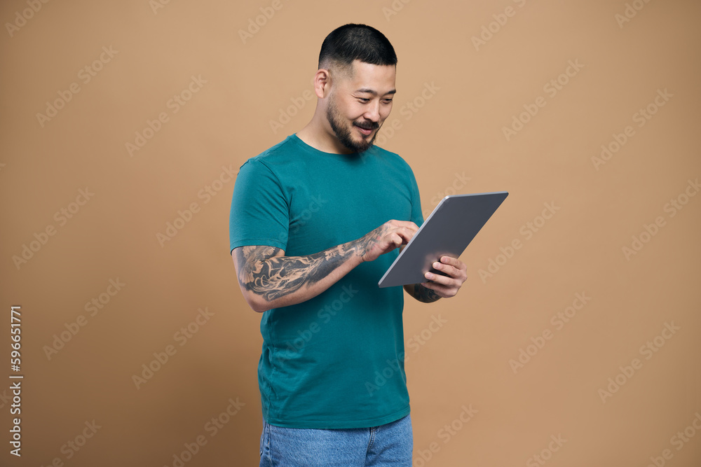 Confident smiling asian man holding modern digital tablet watching videos, communication online isolated on beige background. Happy Korean hipster shopping online, browsing something, ordering food Confident smiling asian man holding modern digital tablet watching videos, communication online isolated on beige background. Happy Korean hipster shopping online, browsing something, ordering food