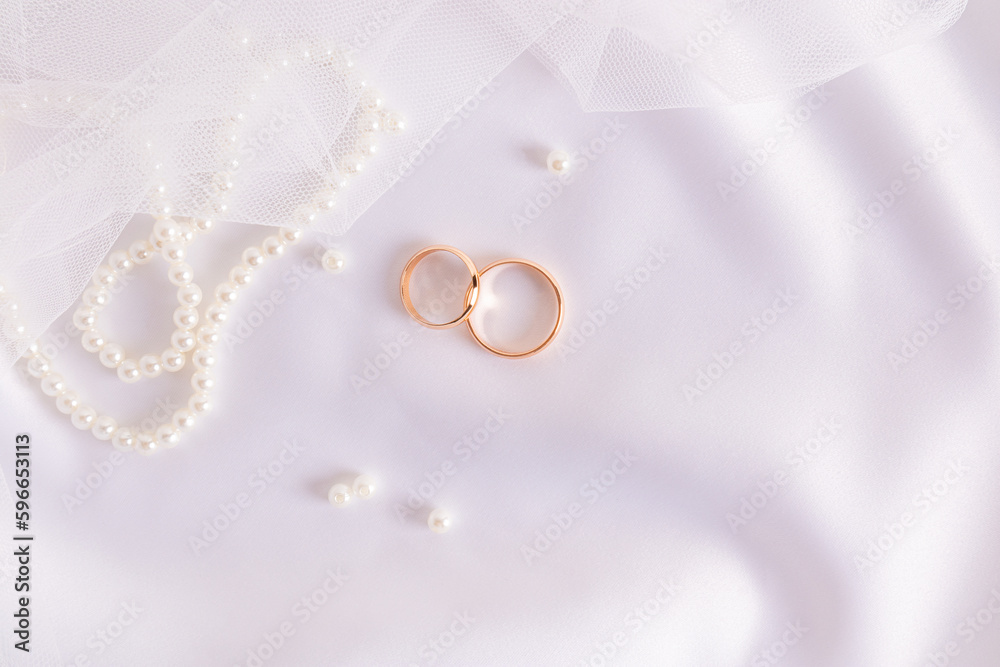 Two gold wedding rings on white satin textiles with pearl beads and veil . Wedding festive background. A copy of the space. Two gold wedding rings on white satin textiles with pearl beads and veil . Wedding festive background. A copy of the space.