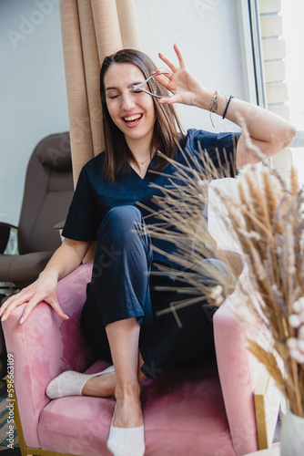 Young laughing European brunette girl manicurist in black uniform sits in a pink chair in a beauty salon and holds manicure scissors to her face