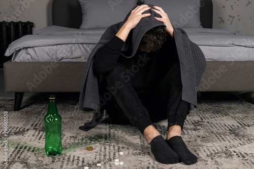 A severely depressed guy, sits on the floor covered with a blanket, leaning down with a bottle and pills. A young man in a hard mental condition. alcohol addiction.