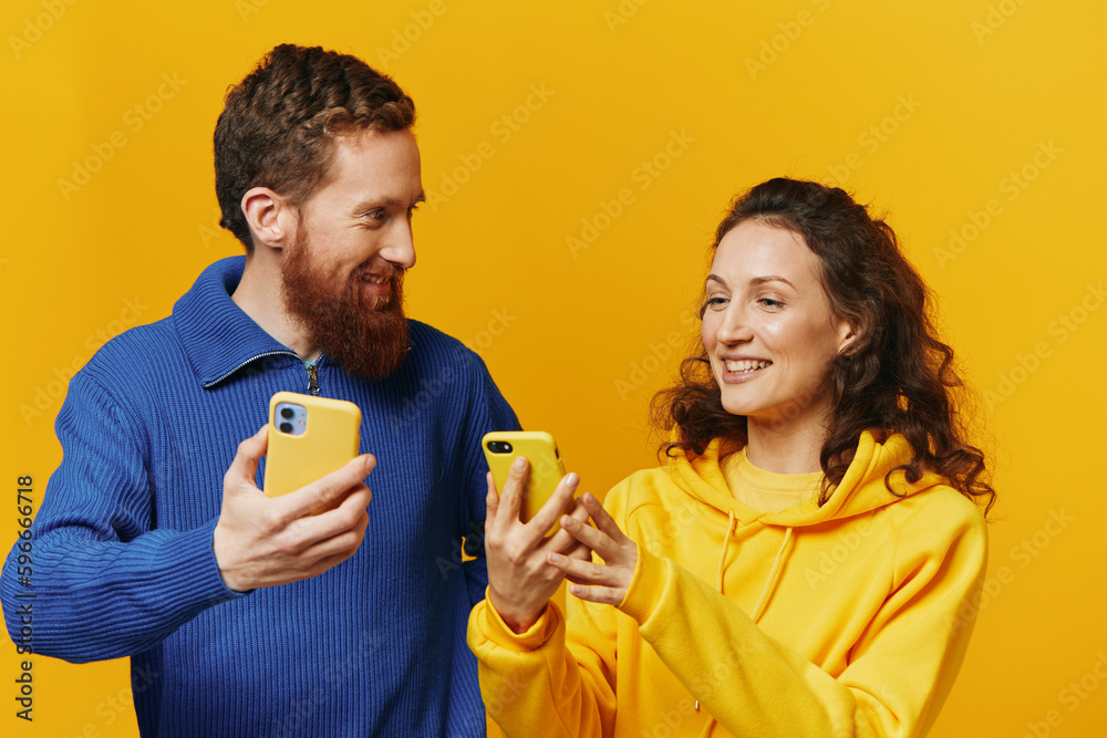 Man and woman couple smiling merrily with phone in hand social media viewing photos and videos, on yellow background, symbols signs and hand gestures, family freelancers. Man and woman couple smiling merrily with phone in hand social media viewing photos and videos, on yellow background, symbols signs and hand gestures, family freelancers.