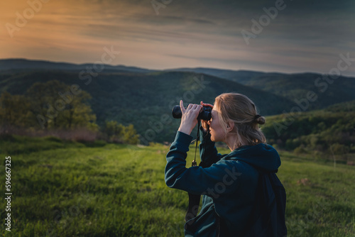 Portrait of a young female tourist looking through binoculars at sunset while walking on green summer mountain valley