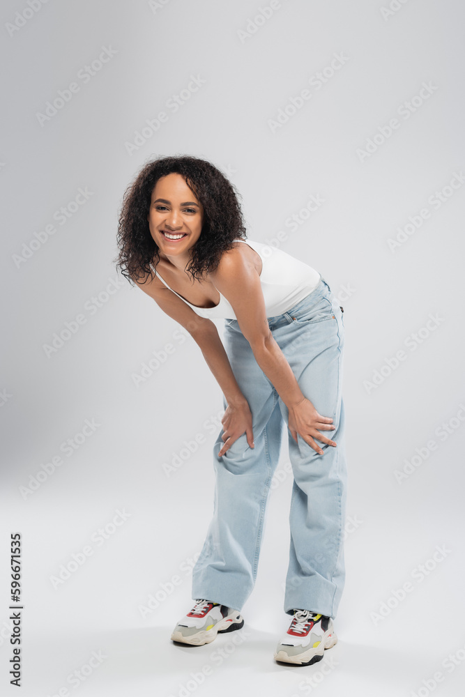 full length of carefree african american woman in blue jeans smiling at camera on grey background. full length of carefree african american woman in blue jeans smiling at camera on grey background.