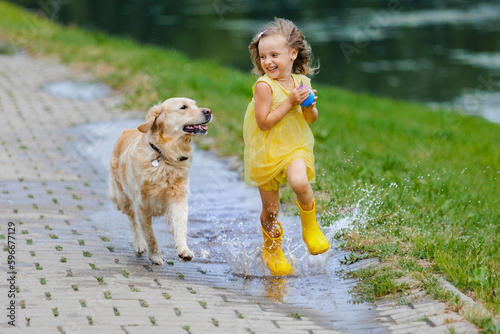 A child in a yellow dress and rubber boots along with her dog, jumps through puddles after the rain.