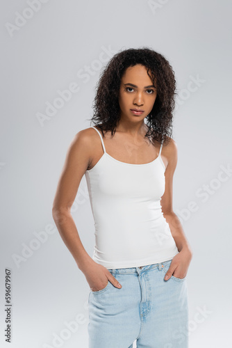 african american woman in tank top posing with hands in pockets of blue jeans isolated on grey.