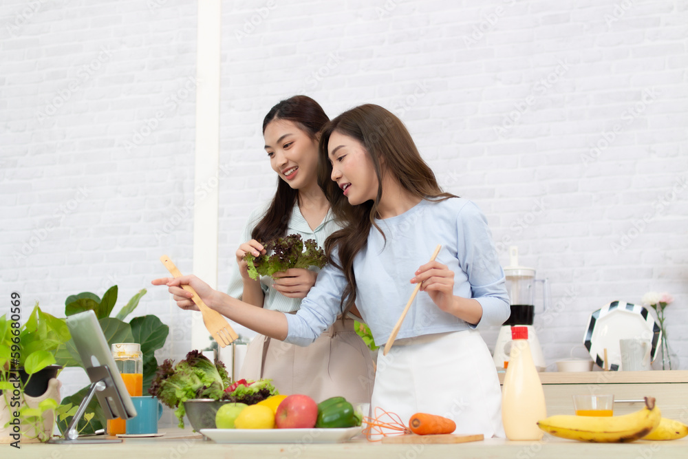 Two young Asian women are reading recipe from the digital tablet in the kitchen. Woman having fun with healthy food salad. Two young Asian women are reading recipe from the digital tablet in the kitchen. Woman having fun with healthy food salad.