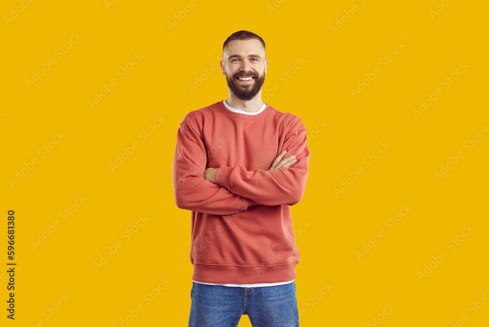 Young smiling bearded man in red sweatshirt and jeans is posing with arms crossed on chest standing isolated on yellow background looking at camera. Sincere human emotions, body language concept. - obrazy, fototapety, plakaty 