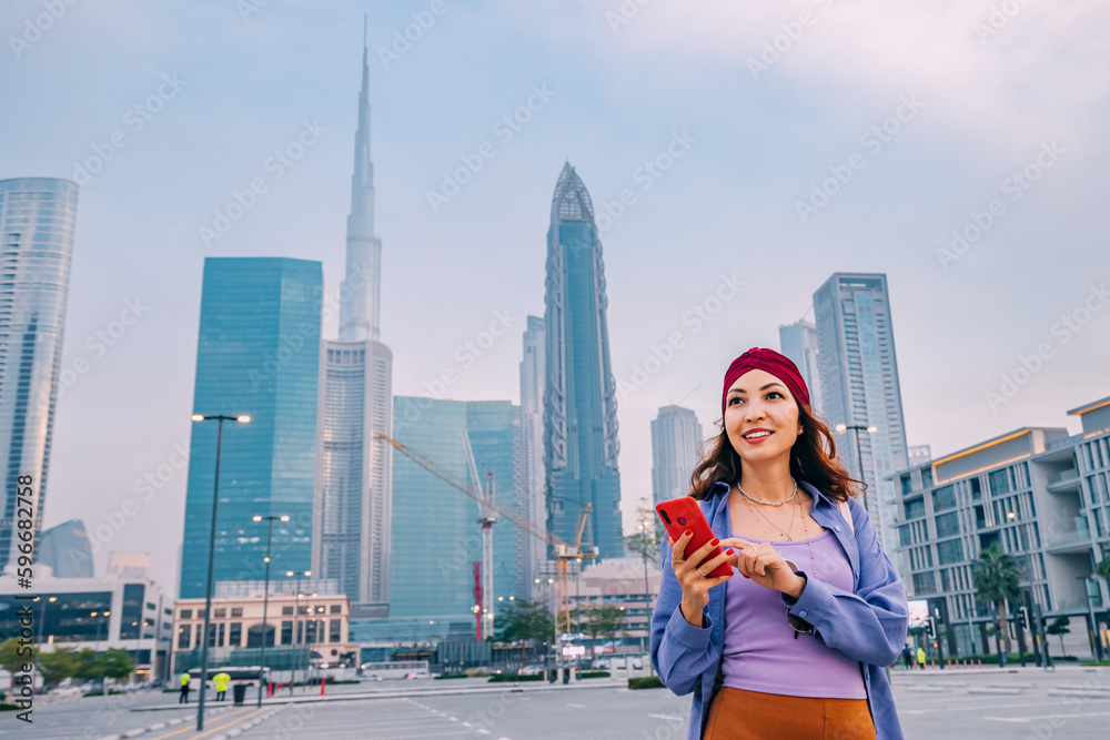 Happy asian girl using smartphone wireless network to connect to internet while walking in Dubai downtown with Burj Khalifa tower and other skyscrapers in background