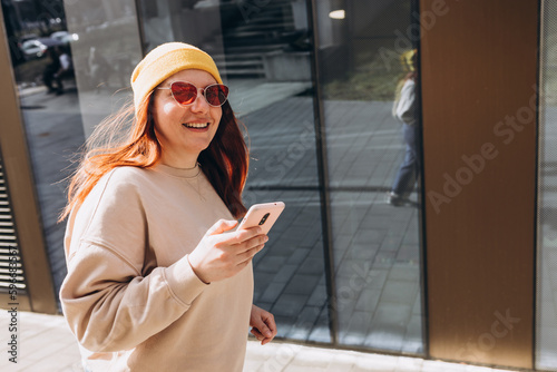 30s girl in yellow hat and pink sunglasses checking social media, happy woman holding smartphone outdoors. Urban lifestyle concept. Traveler © mdyn