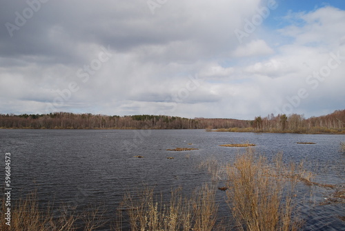 Spring landscape on the lake. Early spring snow almost freed the ground. Trees and bushes are still without snow. Water on the lake without ice. Gloomy sky above with white and gray clouds.