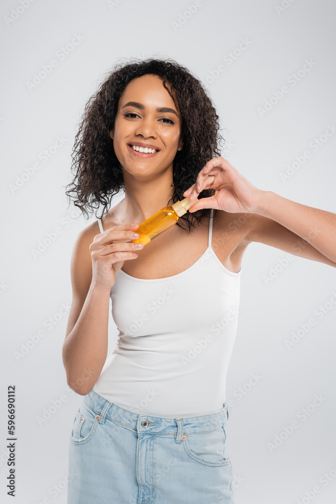 excited african american woman holding bottle of cosmetic oil while looking at camera isolated on grey. excited african american woman holding bottle of cosmetic oil while looking at camera isolated on grey.