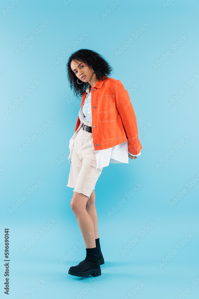 full length of brunette african american woman in orange jacket and black boots looking at camera on blue background.