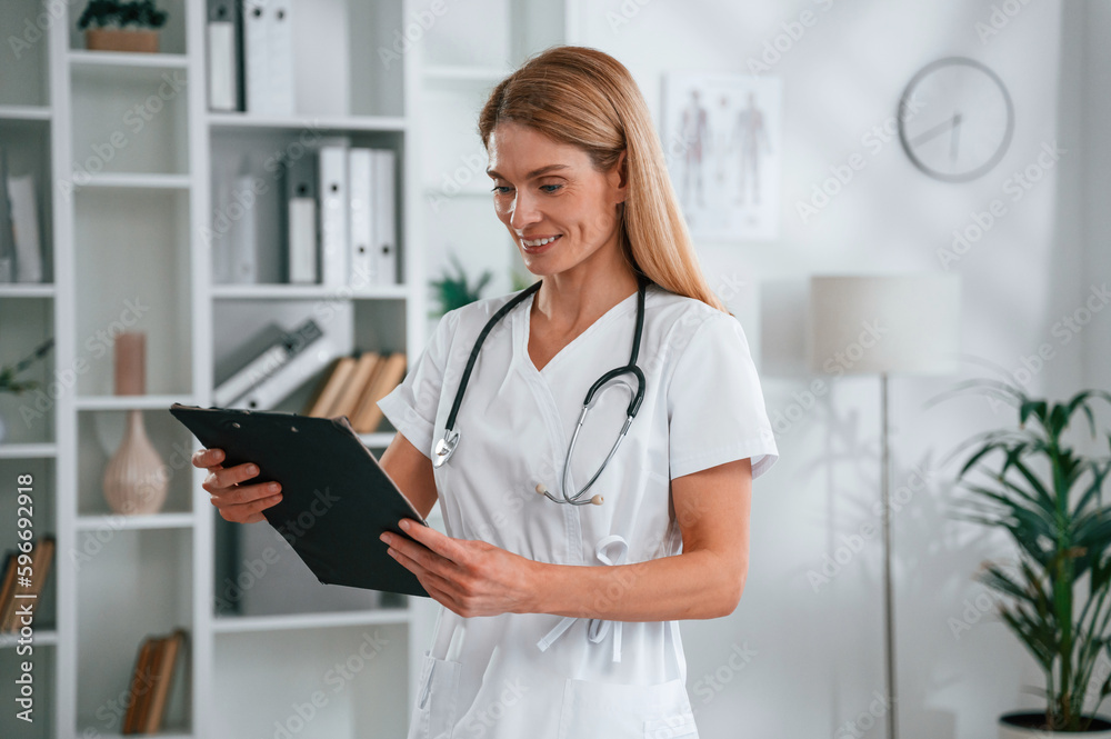 Reading the information in the notepad. Young female doctor in white coat is indoors Reading the information in the notepad. Young female doctor in white coat is indoors