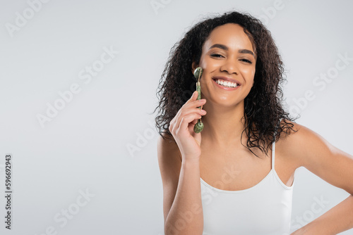 brunette african american woman doing face massage with jade roller isolated on grey.