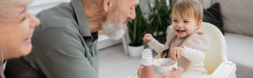 overjoyed girl holding spoons while having breakfast near happy grandparents in kitchen, banner. overjoyed girl holding spoons while having breakfast near happy grandparents in kitchen, banner.