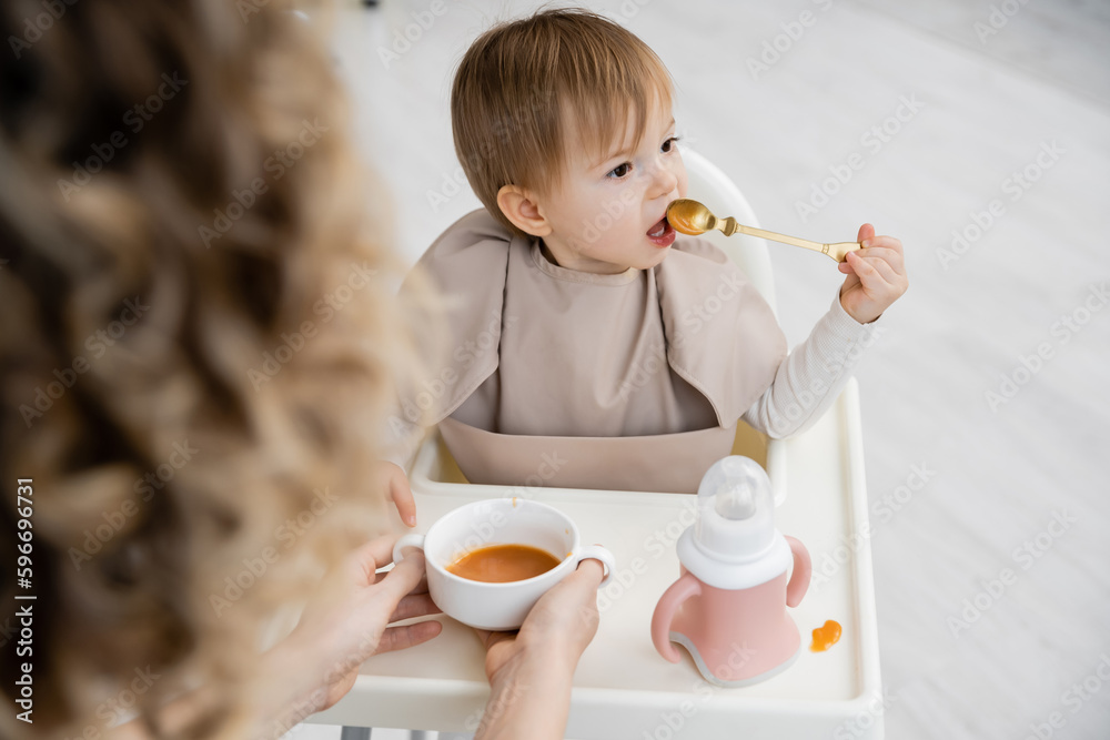 toddler girl sitting on baby chair and eating puree near blurred mom with bowl in kitchen. toddler girl sitting on baby chair and eating puree near blurred mom with bowl in kitchen.