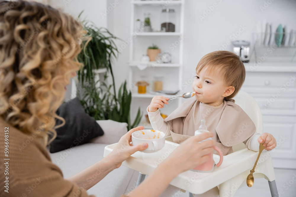 blurred woman holding bowl and baby bottle near toddler daughter eating breakfast in kitchen. blurred woman holding bowl and baby bottle near toddler daughter eating breakfast in kitchen.