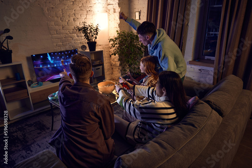 Young people, friends meeting at home in the evening, sitting on couch and playing online sport games, basketball, on console. Concept of friendship, leisure activity, hobby, fun, sport simulator © master1305