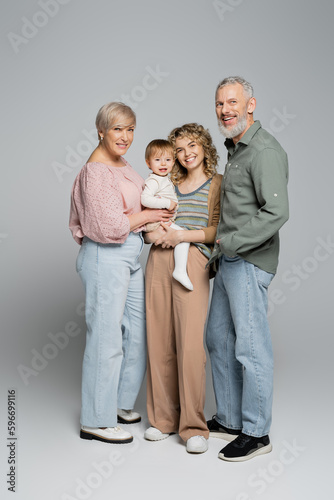 full length of happy middle aged grandparents and adult daughter with baby girl smiling at camera on grey background.