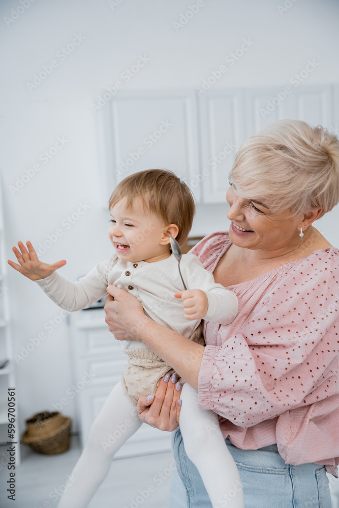 happy mature woman hugging excited granddaughter holding spoon in kitchen.
