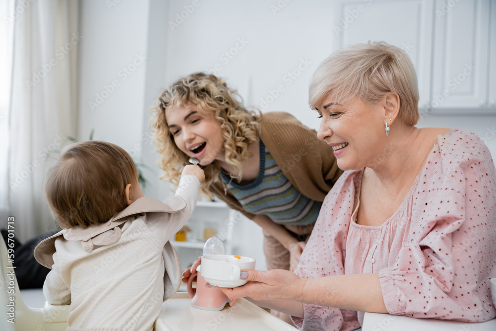 toddler girl feeding mom near smiling grandmother with bowl in kitchen. toddler girl feeding mom near smiling grandmother with bowl in kitchen.