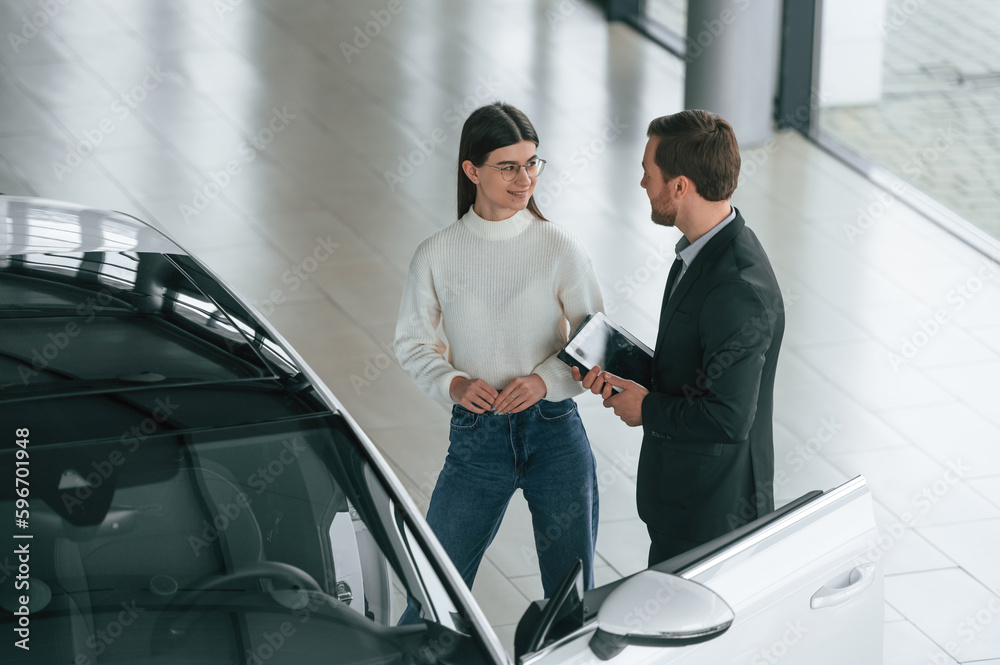 Talking, holding tablet. Man is showing the new automobile to the woman customer in the car dealership Talking, holding tablet. Man is showing the new automobile to the woman customer in the car dealership