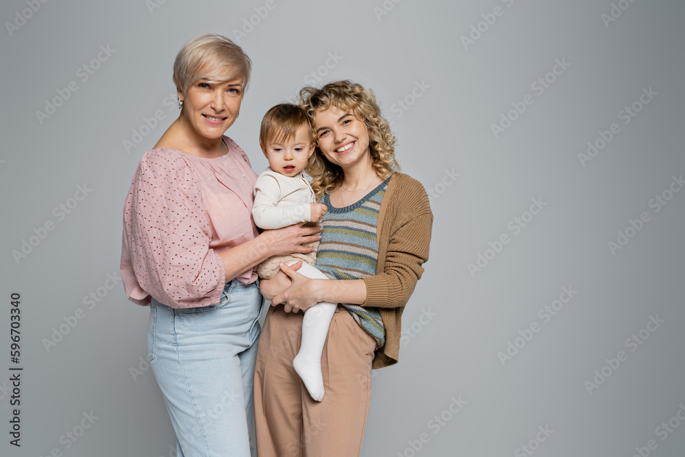 pleased women embracing toddler girl and smiling at camera isolated on grey.