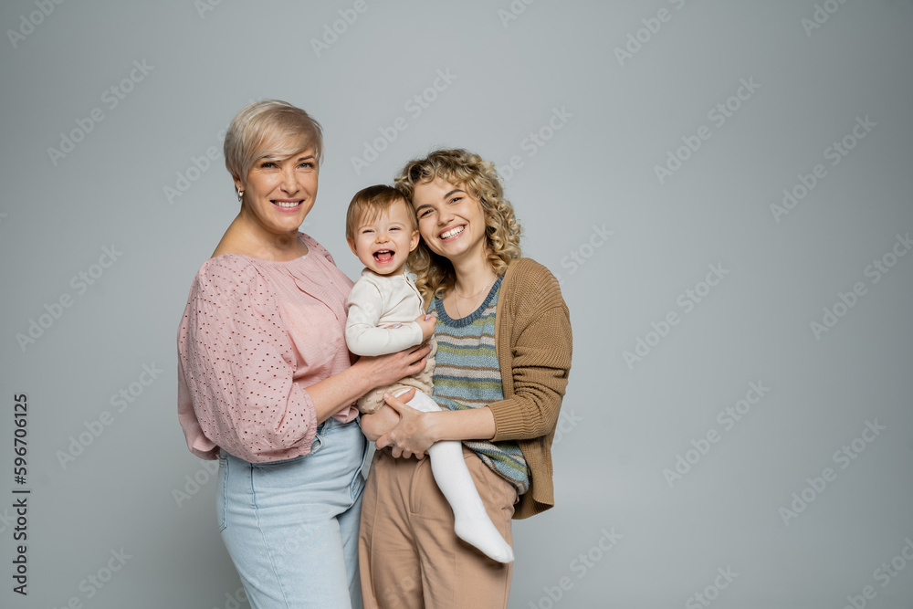 happy women smiling at camera near laughing toddler girl isolated on grey. happy women smiling at camera near laughing toddler girl isolated on grey.