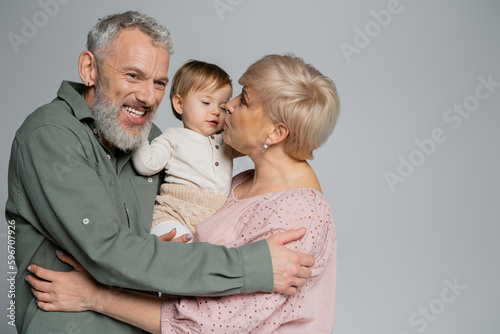 mature woman kissing granddaughter near pleased bearded husband isolated on grey.