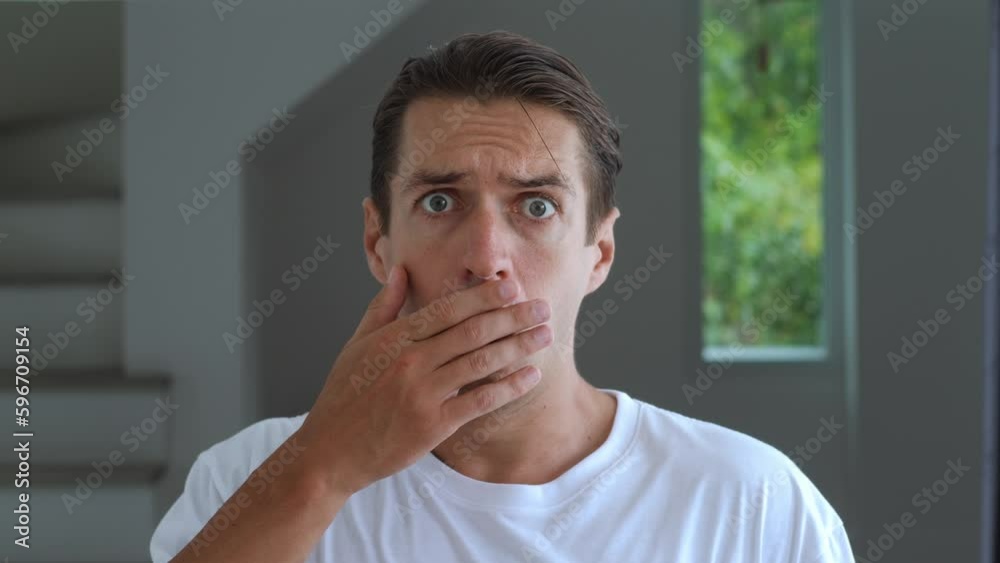 Close-up afraid millennial business man staring eyes and covering mouth in fright reacting on horrible situation on home office apartment background. Portrait of afraid man 30s indoors