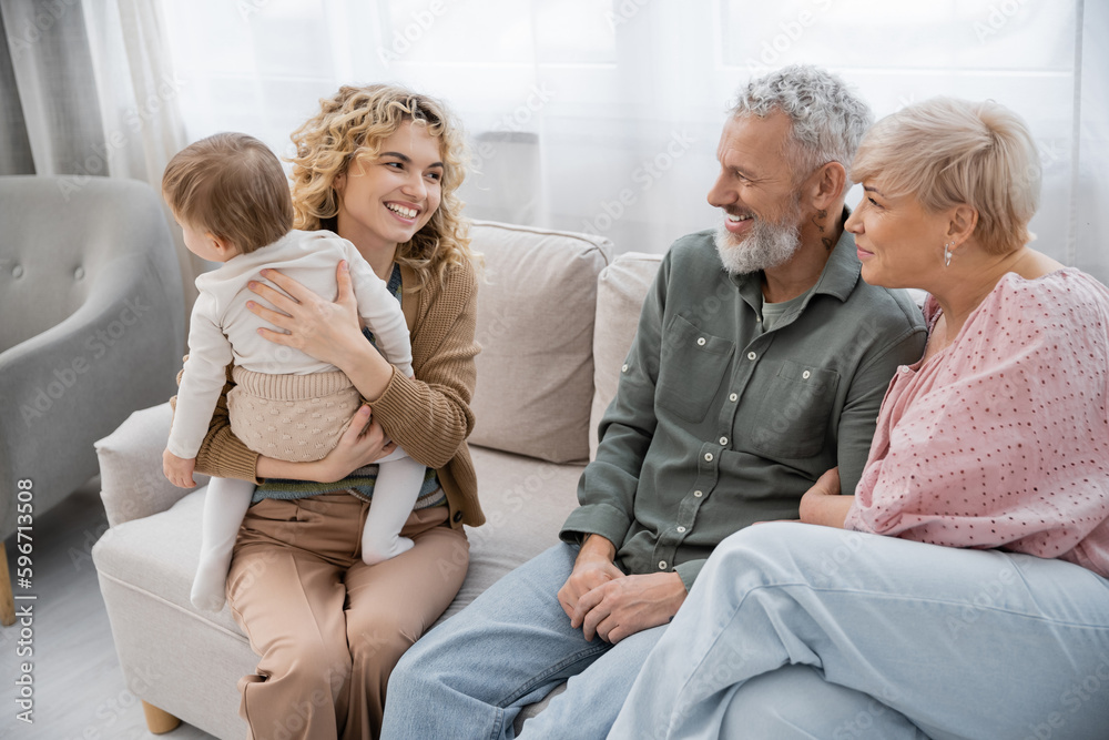 cheerful blonde woman holding baby daughter and looking at happy parents on couch in living room. cheerful blonde woman holding baby daughter and looking at happy parents on couch in living room.