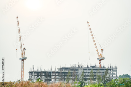 Фотография Skyscraper Under Construction: Towering construction cranes oversee the skeletal frame of a multi-story building emerging against a hazy sky, symbolizing urban development and progress