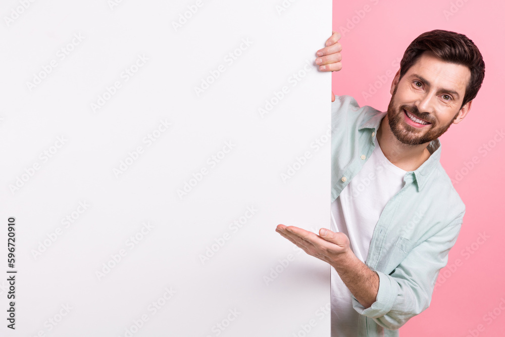 Photo of young funny businessman hiding behind huge placard board demonstrate hand new product name isolated on pink color background