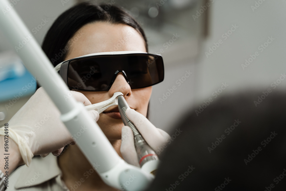 Laser treatment inflammation of the nasal lining, runny nose, sneezing symptoms. Laser treatment of chronic rhinitis. ENT doctor with laser treats girl patient in protective glasses.