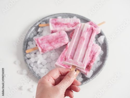 Hand holding the stick of a homemade yogurt and strawberry ice cream popsicle. In the background others on ice in a round silver tray.