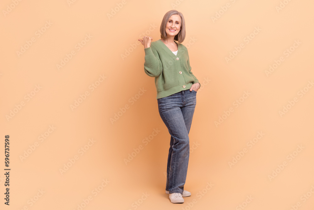 Full length photo of cheerful woman khaki cardigan indicating at discount empty space arm in pocket isolated on beige color background