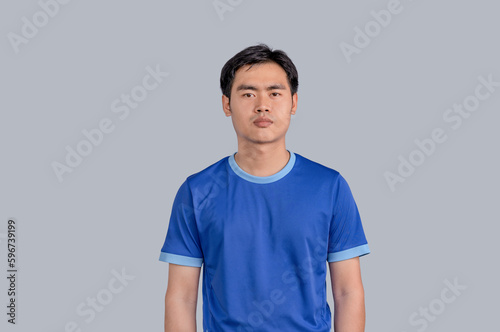 Young Asian man in blue t-shirts standing in studio and serious concentrated face at camera, looking aggressive, isolated on gray background. Relaxed with serious expression on face.
