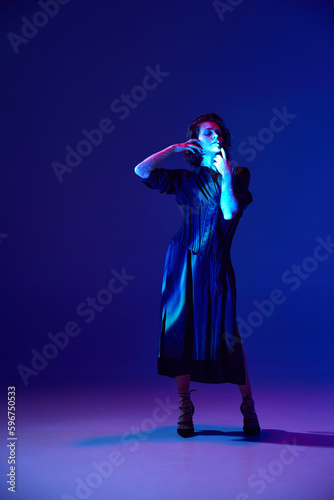 One young sensual woman wearing black long dress touching her face over dark blue studio background in neon light. Tenderness