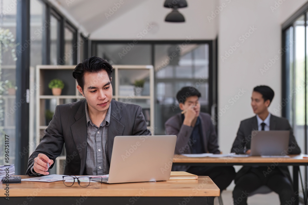 Portrait of confident young businessman using laptop while colleague in background Portrait of confident young businessman using laptop while colleague in background