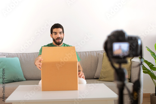 Glad surprised young middle eastern guy blogger at table unpacks cardboard box, shooting video