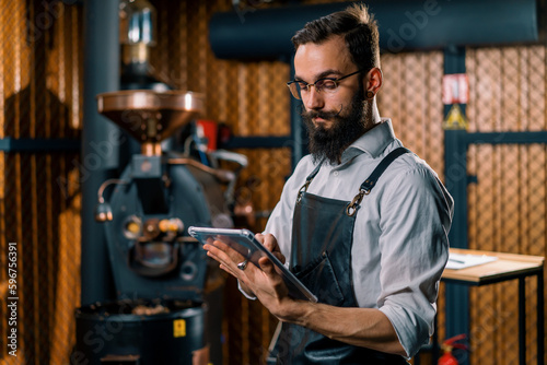serious guy in apron standing in coffee production near machine for roasting coffee beans with tablet in hands