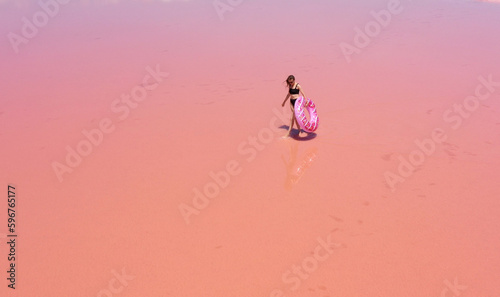 Aerial view. Young woman in black swimsuit carries an inflatable pink float in her hands have fun dancing on the pink salt lake. Concept summer festivals, holiday, travel vacation, freedom, sun, enjoy