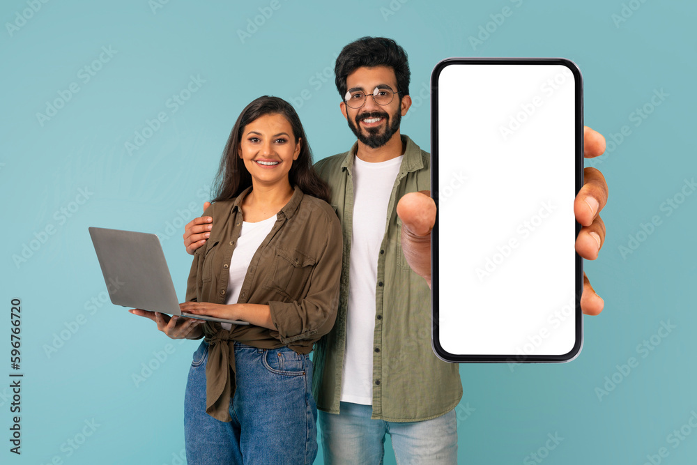 Positive mixed race couple using laptop, showing big phone, mockup Positive mixed race couple using laptop, showing big phone, mockup