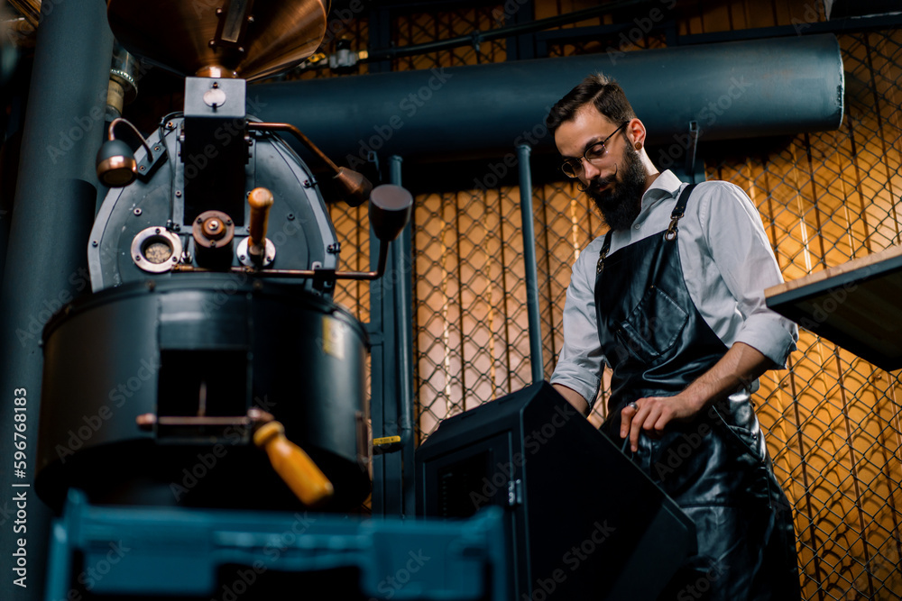 a handsome young bearded man in an apron enters data into a machine for roasting coffee beans at a factory