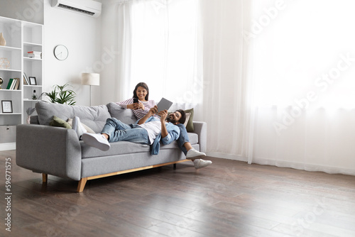 Candid shot of eastern couple chilling on couch, using gadgets