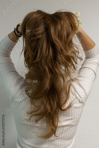 Young woman holding her natural beautiful long wavy hair from behind, model in a white blouse on a white wall, photo in a movement