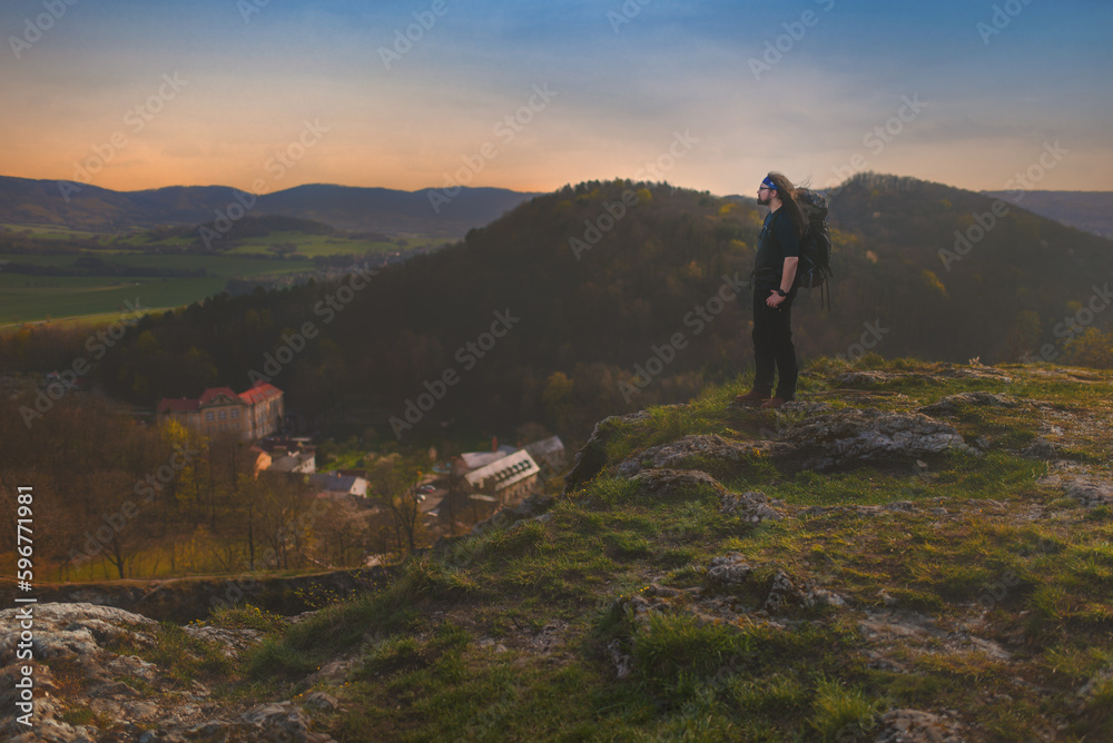 Obraz premium A lone longhaired man with a backpack on his back is standing on a sunny rocky lookout above the town of Štramberk in the Podbeskydí Nature Park.